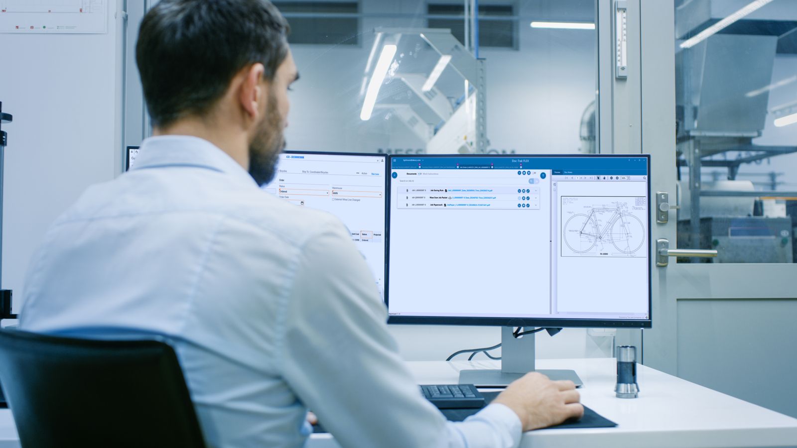 An engineer at two monitors with a drawing open beside the SyteLine record