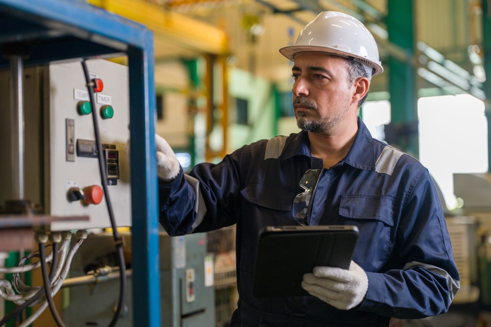 A technician in a hard hat reading a machine panel with a tablet in hand